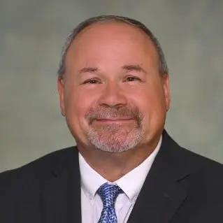 Professional headshot of a smiling middle-aged man in a dark suit and blue tie against a blurred neutral backdrop.